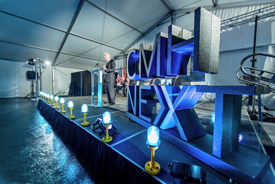 Joseph Nardone stands at a podium on stage, addressing a crowd. Lined up on the stage are several runway lights that are illuminating the area in blue. On the right side of the stage is a large CMH Next statue.